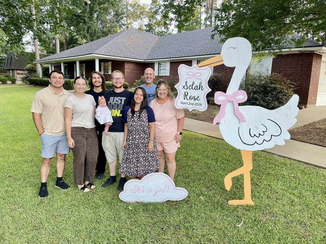A family gathered in their front yard around a white wooden girl stork with a pink bow, a personalized name plate, and an &ldquo;It&rsquo;s a girl!&rdquo; cloud sign