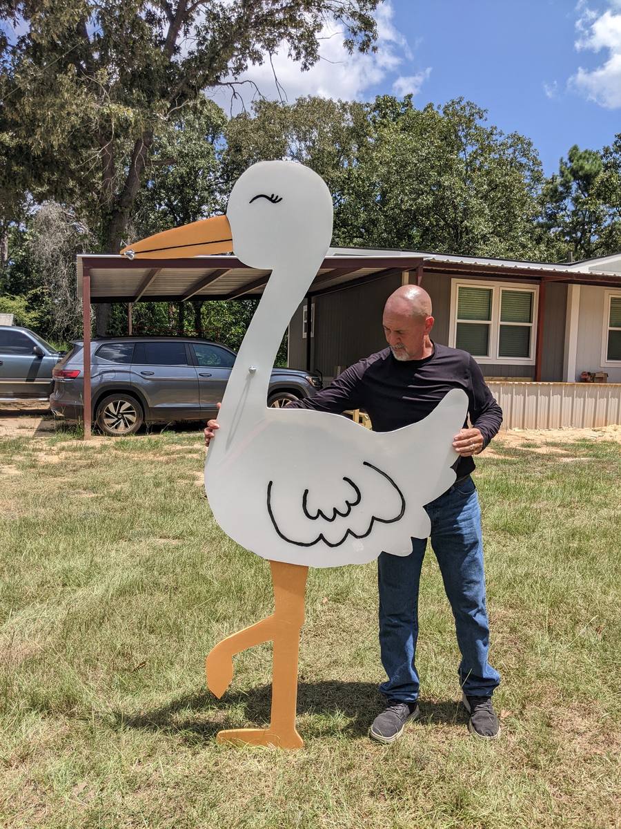 A man carrying a tall white wooden stork across a grassy front yard to stand it up in place