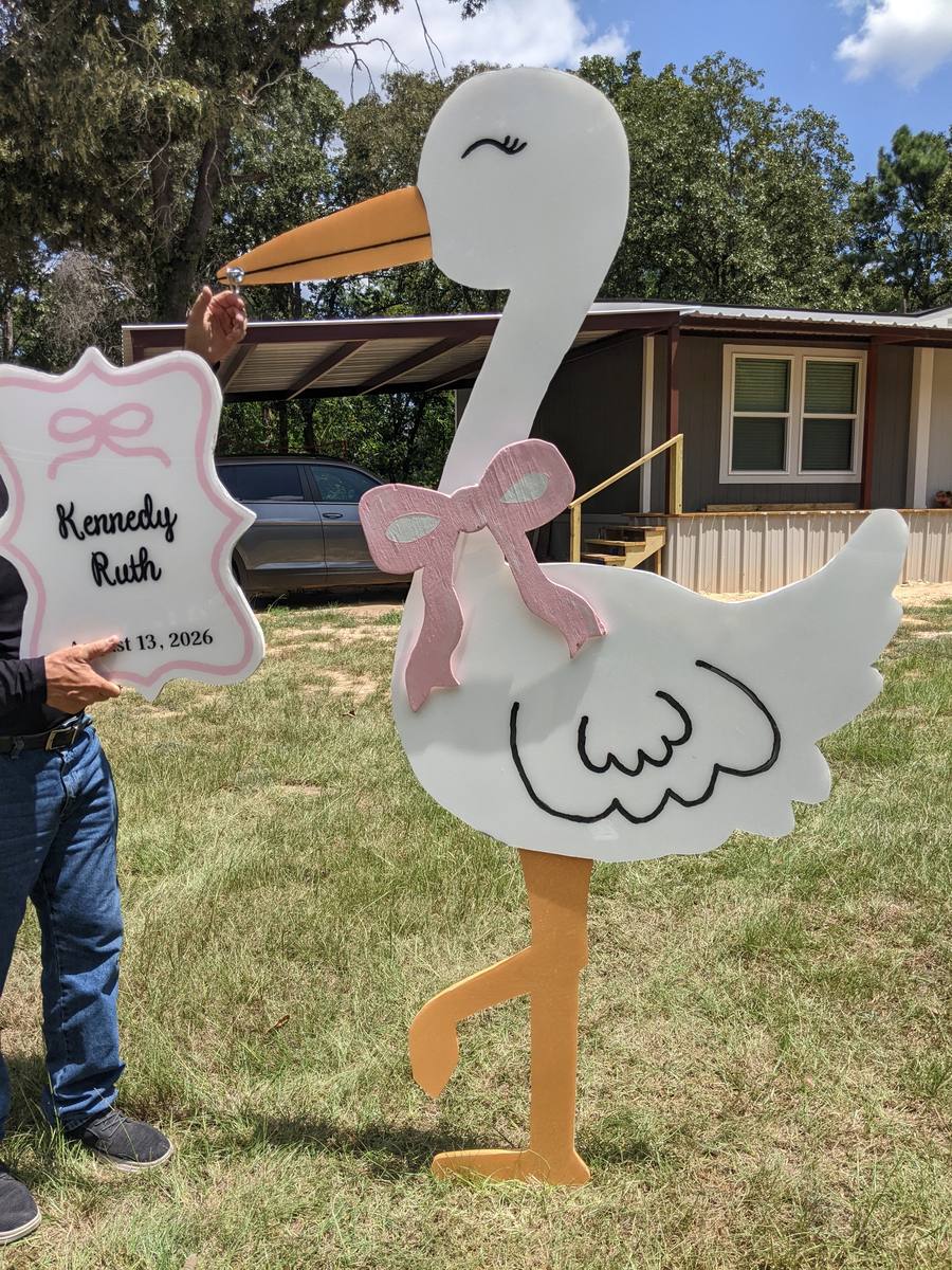 A pink bow being added to the stork while a personalized name plate reading &ldquo;Kennedy Ruth, August 13, 2026&rdquo; is held up beside it