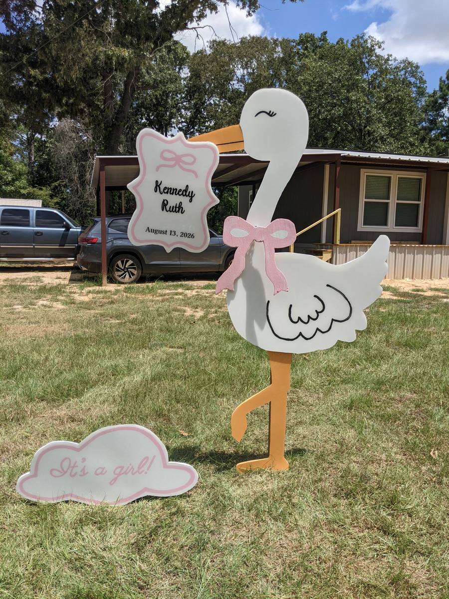 The finished girl stork display in a front yard with a pink bow, a &ldquo;Kennedy Ruth, August 13, 2026&rdquo; name plate, and an &ldquo;It&rsquo;s a girl!&rdquo; cloud sign in the grass